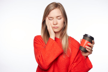 Sleepy attractive blond Caucasian female student worked all day at diploma, poses indoors with take away cup of coffee, wants to have rest, being tired and exhausted. Woman in red sweater closes eyes.