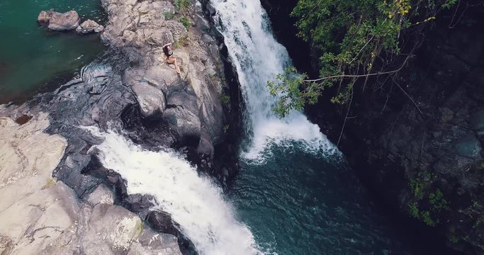 Aerial drone view of young beautiful tourist sitting near the Kroya waterfall of the Bali island, Indonesia. Having fun in the wild nature