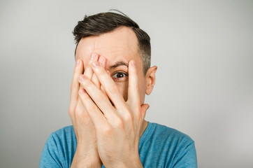 A young guy dressed in a blue t-shirt covers his face with his hands, isolated against a light background.