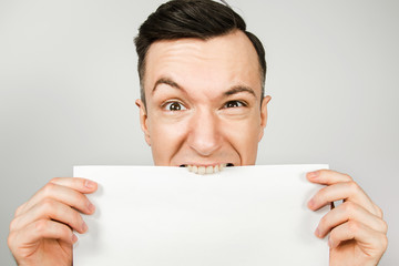 Young guy dressed in a blue t-shirt holds blank of A4 paper and bites him, isolated on a light background. Close-up portrait.
