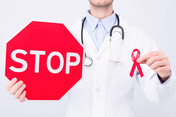 Oncological disease concept. Doctor wearing white coat and stethoscope holding red ribbon as a symbol of aids and stop sign