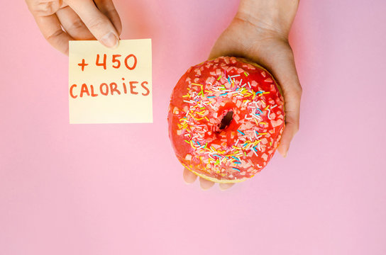Woman hand holding pink donut with sprinkles extra calories fat fast food excess weight unhealthy on yellow background