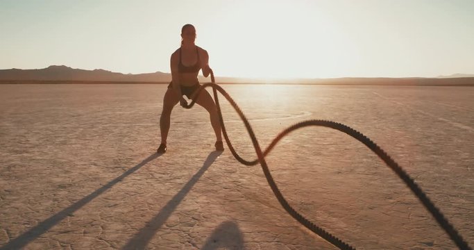Slow motion: Athletic woman doing crossfit battle rope workout outside at sunset in desert 
