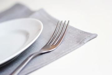 Empty white plate on a gray napkin with fork on white table. Table setting, preparation for food.