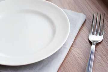 Empty white plate on a gray napkin with fork and glass on brown table. Table setting, preparation for meals.