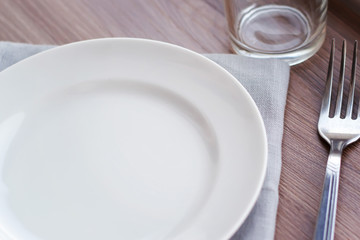 Empty white plate on a gray napkin with fork and glass on brown table. Table setting, preparation for meals.