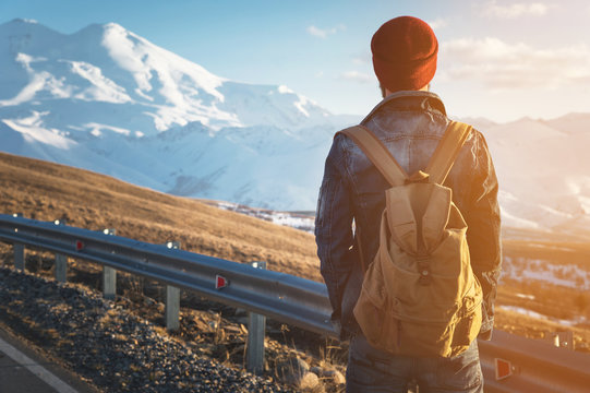 Bearded tourist hipster man in sunglasses with a backpack stand back on a roadside bump and watching the sunset against the background of a snow capped mountain - Powered by Adobe