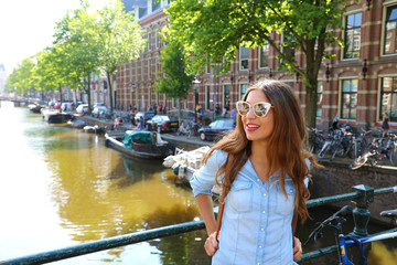 Happy young woman with sunglasses standing on a bridge looking to the side with her bike enjoying morning air on Amsterdam canal, Netherlands - obrazy, fototapety, plakaty