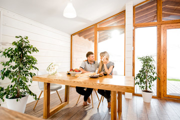 Fototapeta na wymiar Young and happy couple having a breakfast sitting at the wooden table in the modern country house with big window on the background