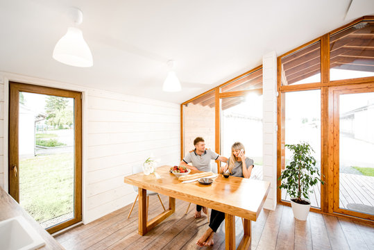 Young and happy couple having a breakfast sitting at the wooden table in the modern country house with big window on the background