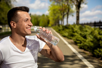 Profile of smiling guy drinking water from bottle. He is spending sunny day in green park. Enjoying warm weather walking outside concept