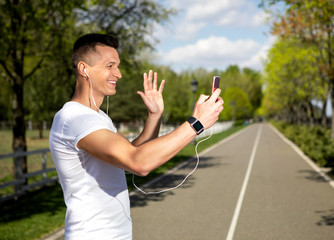 Profile of cheerful male waving hello to smartphone. He is speaking with somebody using earphones and camera for online connection. Guy is spending day in countryside and wearing smartwatch