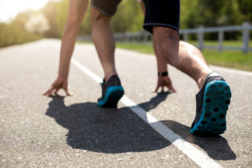 Close-up of male feet in crouch start on asphalt track outside and turning back to camera. He is bending body and putting hands on road while feeling concentrated. Guy is wearing trainers and watch