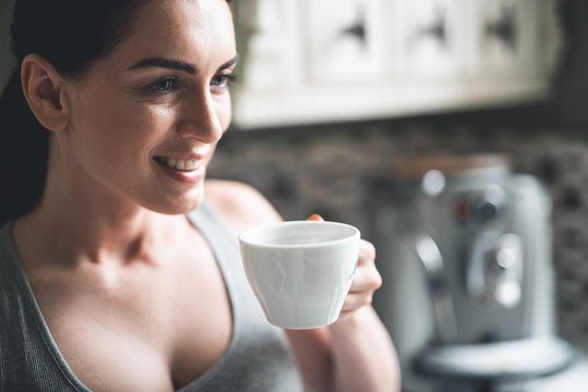 Close up on joyful female holding cup in hands and smiling. She is drinking her favorite drink and admiring free time in kitchen