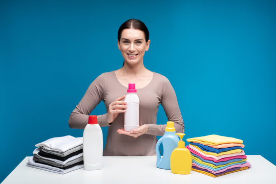 Waist up portrait of smiling female isolated on blue background. She is holding softener in hands. Delightful woman is standing at table with liquids and piles of clean clothes