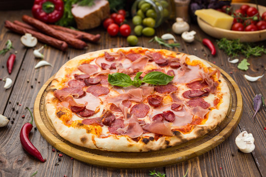 Delicious italian pizza with ham, bavarian sausages and prosciutto decorated by basil leaves on wooden background