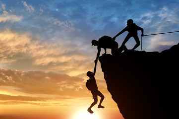 Silhouette of three male climbers rescuing another male climber pulling his arm