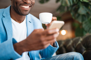 Beaming male using mobile while tasting cup of coffee. Glad guy during leisure concept. Low angle