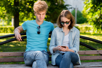 A young guy with a girl is sitting on a bench. In summer in the park in nature. Hands holding a phone communicates in social networks. Dressed in casual clothes. Sunglasses.