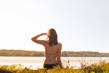 Beautiful slim woman enjoying a drink near lake at sunset of warm sunny afternoon. Young fit female drinking lemonade at river bank