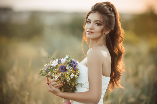 Pretty young bride with bouquet of wildflowers in field over sunset lights