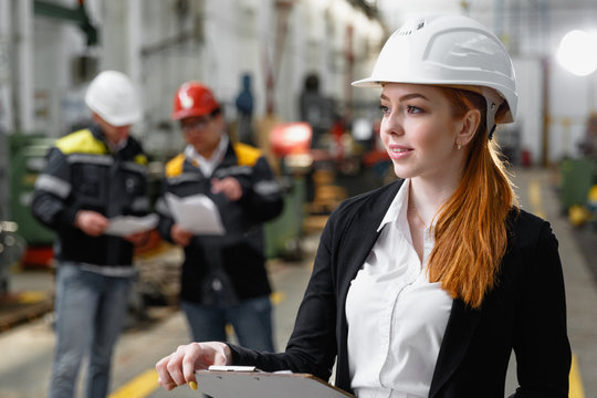 A successful engineer girl checks the technical documentation at the plant. In the background, two workers in helmets and working uniforms.