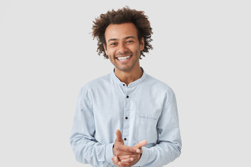 Waist up portrait of cheerful handsome African American male keeps hands together, smiles broadly, dressed in elegant shirt, being in high spirit after date with girlfriend, shares impressions