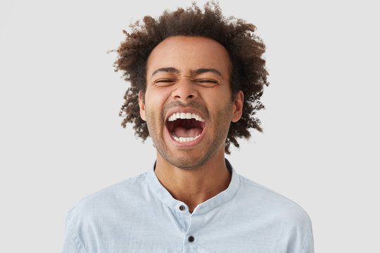 Photo of overjoyed dark skinned mixed race male with curly hairstyle, laughs happily, keeps mouth widely opened, shows white teeth, can`t stop giggling, hears anecdote, has funny expression.
