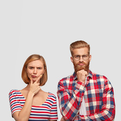 Thoughtful young groupmates try to find solution, have pensive clever expressions, hold chins, look seriously into camera. Attractive red haired guy with ginger beard collaborates with pretty female