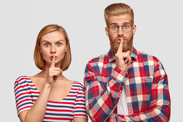 Studio shot of young European boyfriend and girlfriend, show silence sign, looks with surprised expressions, demonstrate shush gesture, stand close to each other, isolated over white background