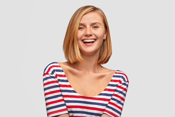 Fototapeta premium It`s awesome! Positive Caucasian female with broad smile, shows white teeth, laughs at good joke, likes funny story from interlocutor, has slim body, dressed in striped jacket. Positive emotions