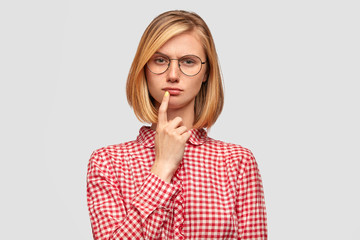Indoor shot of serious thoughtful female student has clever expression, keeps fore finger near mouth, tries to remember material she learned, wears round glasses stylish blouse, isolated on white wall