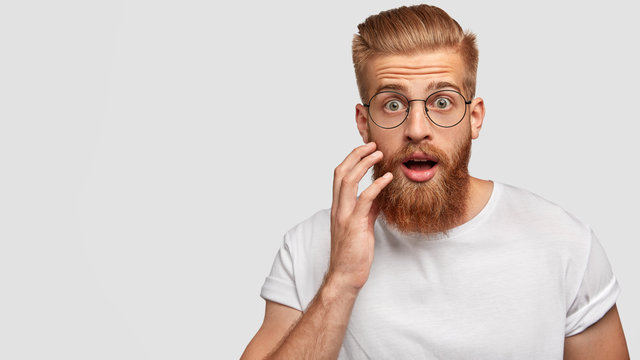 Astonished attractive male has ginger long beard, wonders sudden news, keeps mouth slightly opened, stares at camera, wears casual clothes and spectacles, poses against white wall with blank space