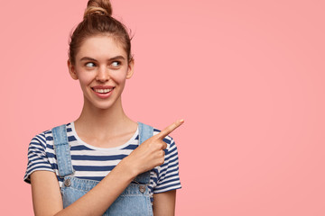 Obraz premium Cheerful Caucasian female with hair bun, dressed in casual outfit, shows blank copy space for your advertisement or promotional text, isolated over pink background. People and happiness concept