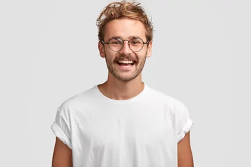 Fotobehang Horizontal shot of happy hipster male with toothy smile, wears casual white t shirt and glasses, being in good mood after unforgettable journey with girlfriend, isolated on white background. © Wayhome Studio