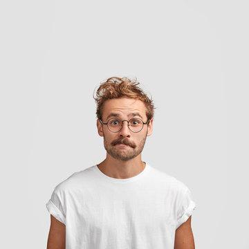 Vertical shot of puzzled European male with curly haircut, purses lips and looks in bewilderment, surprised to see big sales, dressed in casual white t shirt, isolated over studio background