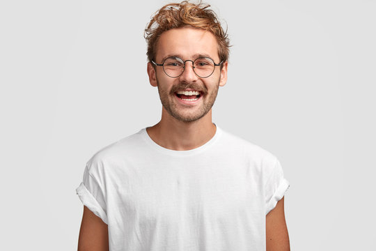 Horizontal shot of happy hipster male with toothy smile, wears casual white t shirt and glasses, being in good mood after unforgettable journey with girlfriend, isolated on white background.