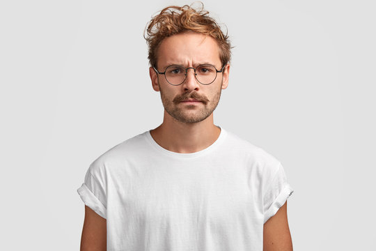 Indoor shot of serious man with grumpy expression, being discontent with noisy neighbours, dressed in casual white t shirt and spectacles, poses indoor. Attractive hipster has strict look and mustache
