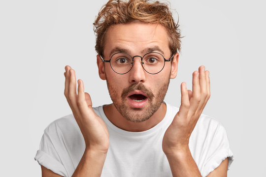 Embarrassed attractive young male clasps hands near head, keeps mouth slightly opened, being surprised of what he sees, has ginger mustache, dressed casually, isolated over white background.
