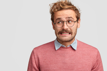 Puzzled attractive bearded emotive male wonk in spectacles, looks with worried embarrassed...