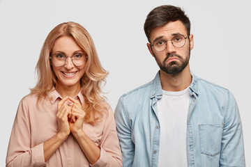 Partnership and different emotions concept. Positive middle aged blonde female in eyewear keeps hands together, smiles positively, stands near his young male partner who frowns face in discontent