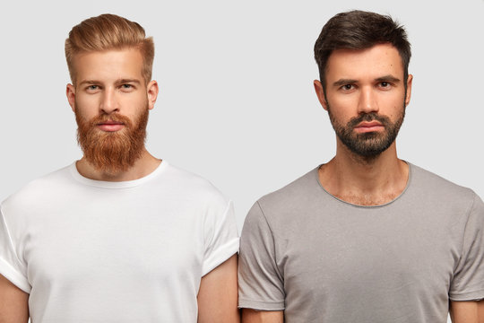 Bearded serious male friends with trendy haircut, stand close to each other, think where spend free time, dressed casually. Two stylish young men colleagues collaborate for creating common project