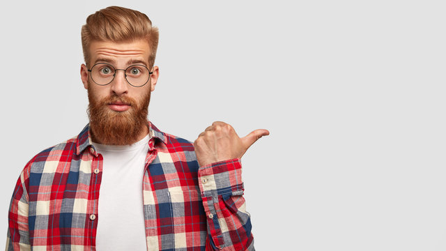 Fashionable male hipster with trendy haircut and ginger thick beard, dressed in checkered shirt, has stunned expression, points with thumb aside, shows blank space for your advertising content