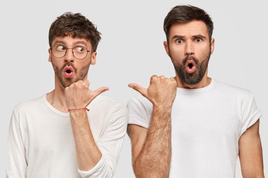 Photo of two amazed guys indicate at each other and have stupefied expressions, keep jaws dropped, dressed in casual white t shirts in one tone with background. People, emotions, gestures concept