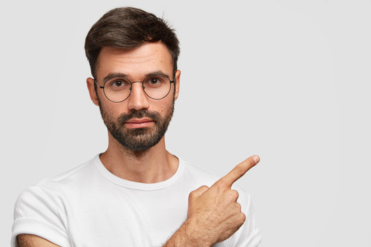 Horizontal shot of attractive European male with serious expresssion, wears round glasses, shows blank space for your advertising content, suggests you to go in this place. People and advertisement