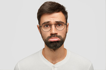 Sullen unshaven male with displeased expression, curves lower lip, has miserable expression, being...
