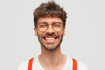 Positive bearded male hipster smiles broadly, has pleased expression, laughs at something funny,...