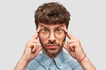 Serious bearded young Caucasian male keeps fingers on temples, tries to recollect something important, looks stressfully, focused on information, thinks over suggestion, isolated on white wall