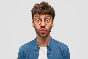 Emotive handsome young guy pouts lips with wonderment, stares through big glasses, has dark...