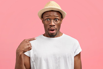 Amazed handsome dark skinned male tourist in straw hat and white t shirt, points at himself with...